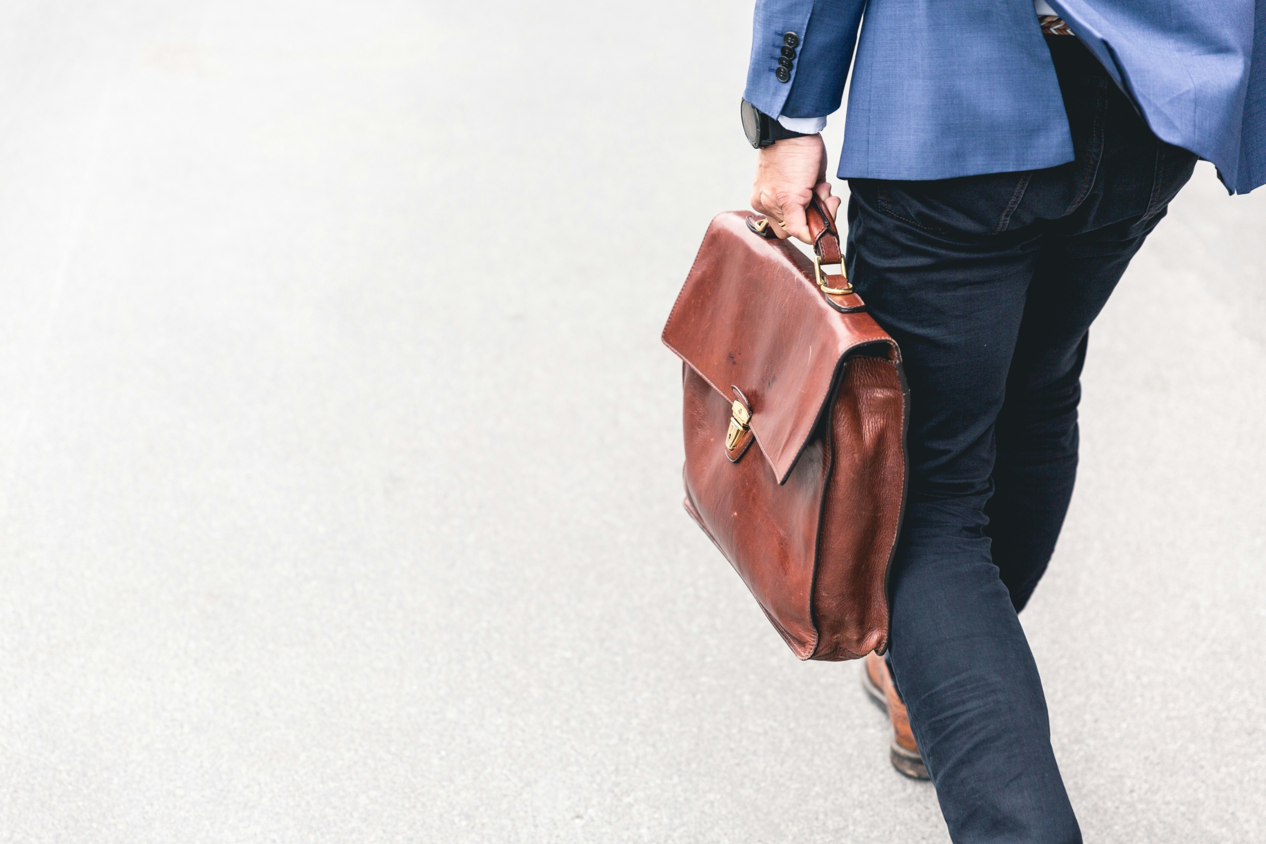 A man walks across a sidewalk, holding a brown leather suitcase and wearing a blue suit jacket
